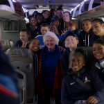 Mae's farewell speech in Ted Lasso S4E9 — Mae beaming centre-frame surrounded by the full Lady Greyhounds squad on the bus to Bristol