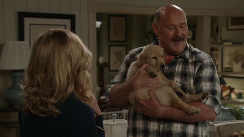 A smiling man in a plaid shirt cradles a golden retriever puppy while a blonde woman, seen from behind, watches in the living room, in Georgie & Mandy’s First Marriage.