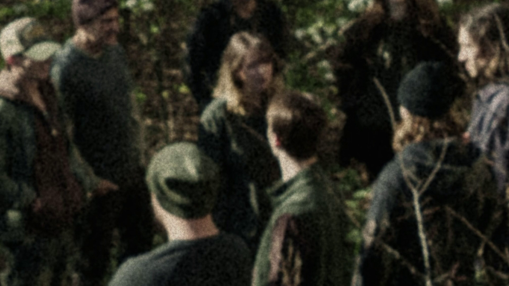 A grainy, out-of-focus group of people in beanies and caps standing close together among bare branches in the woods, in a still from Cupertino.
