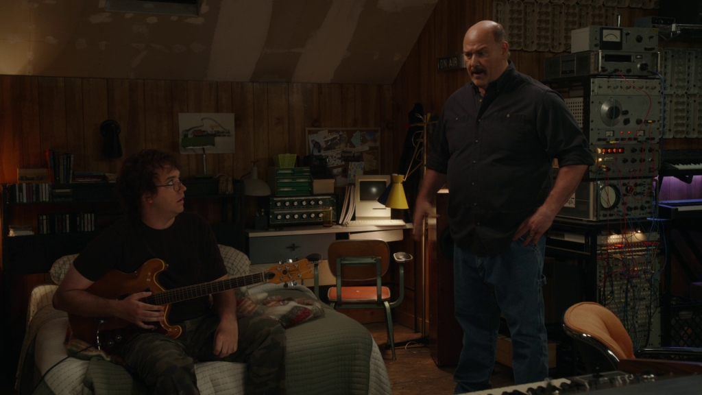 Connor sits on his bed holding a guitar while Jim stands over him in a cluttered attic studio full of synthesizer gear.