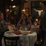 Mary and Fagenbacher at their restaurant table while Georgie stands beside them, looking down at the date he’s about to interrupt.