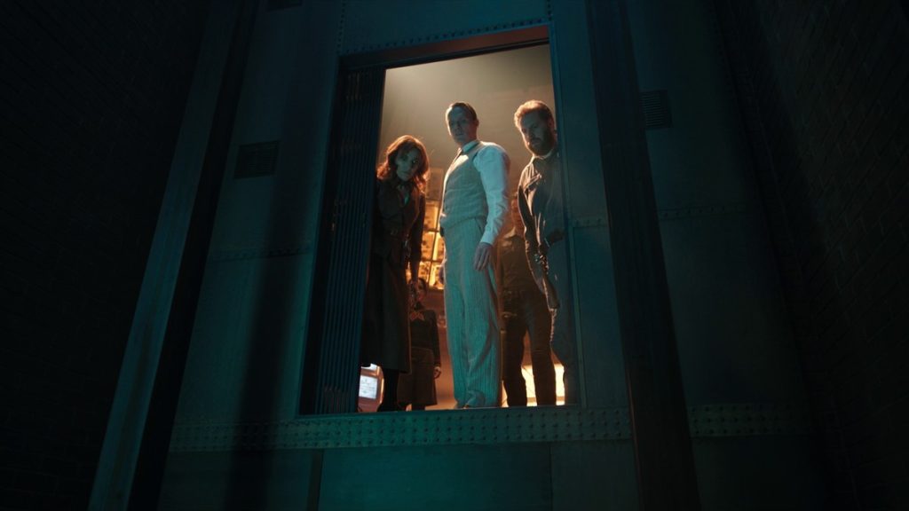 Vision in a white pinstripe suit stands with Friday and U, the bearded household AI, looking down from a lit doorway in a dark chamber in VisionQuest